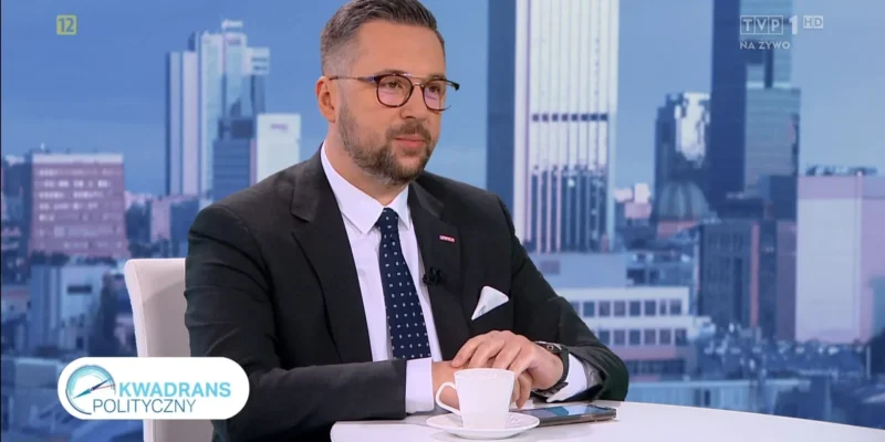 Mężczyzna w garniturze i okularach siedzi w studiu telewizyjnym, na stole ma filiżankę kawy i telefon, w tle widać panoramę miasta oraz logo programu „Kwadrans Polityczny”.