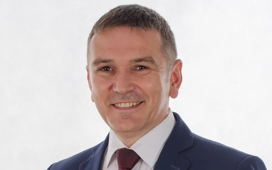 Portret mężczyzny w granatowym garniturze i bordowym krawacie na jasnym tle, uśmiechnięty.