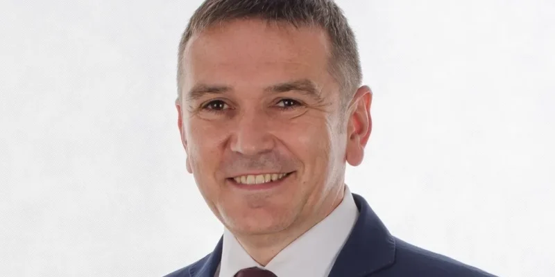 Portret mężczyzny w granatowym garniturze i bordowym krawacie na jasnym tle, uśmiechnięty.