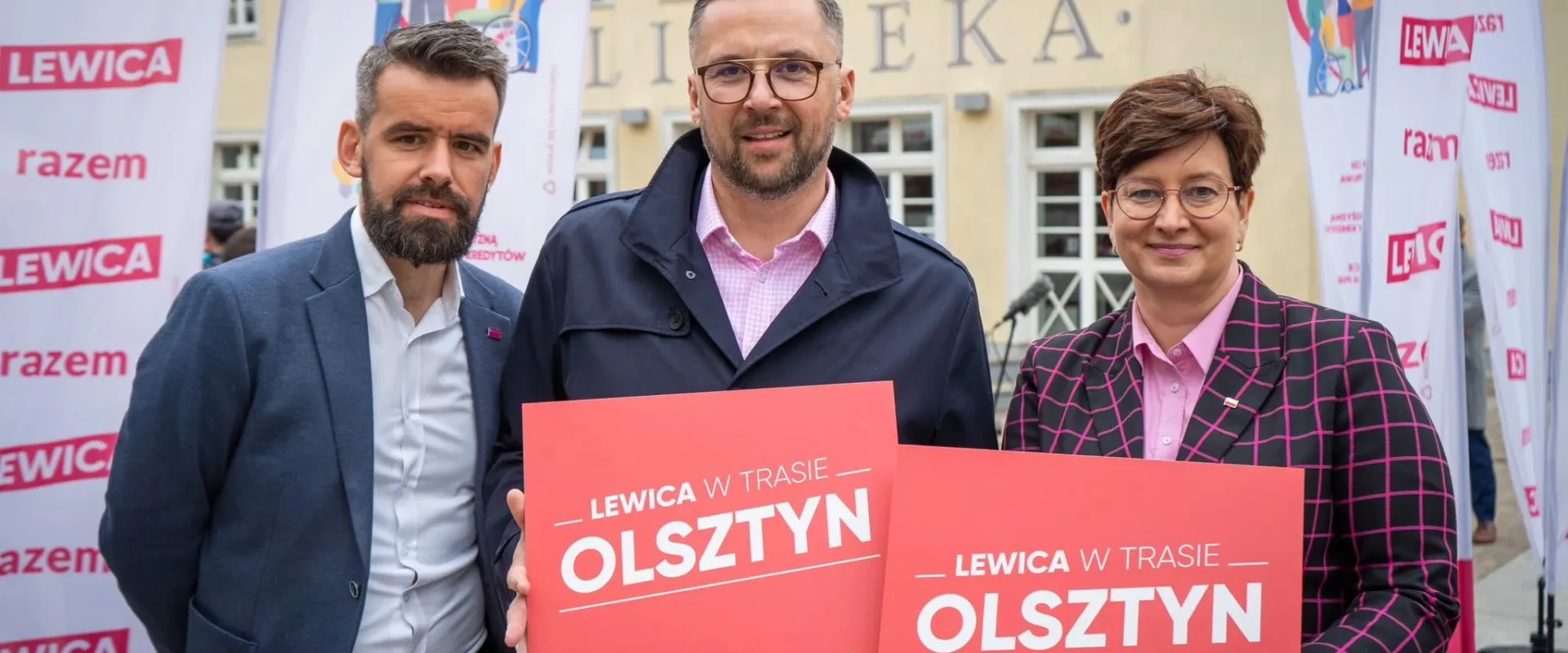 Trzy osoby stoją przed budynkiem otoczone flagami Lewicy, trzymając czerwone kartki z napisem „LEWICA W TRASIE OLSZTYN”.