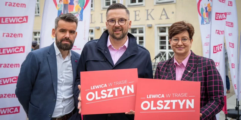 Trzy osoby stoją przed budynkiem otoczone flagami Lewicy, trzymając czerwone kartki z napisem „LEWICA W TRASIE OLSZTYN”.