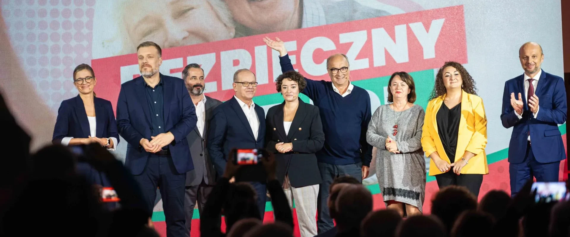 Grupa osób stojących na scenie podczas konwencji Bezpieczny Senior, na tle dużego kolorowego banera z napisem BEZPIECZNY i logo Lewicy.