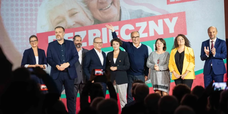 Grupa osób stojących na scenie podczas konwencji Bezpieczny Senior, na tle dużego kolorowego banera z napisem BEZPIECZNY i logo Lewicy.