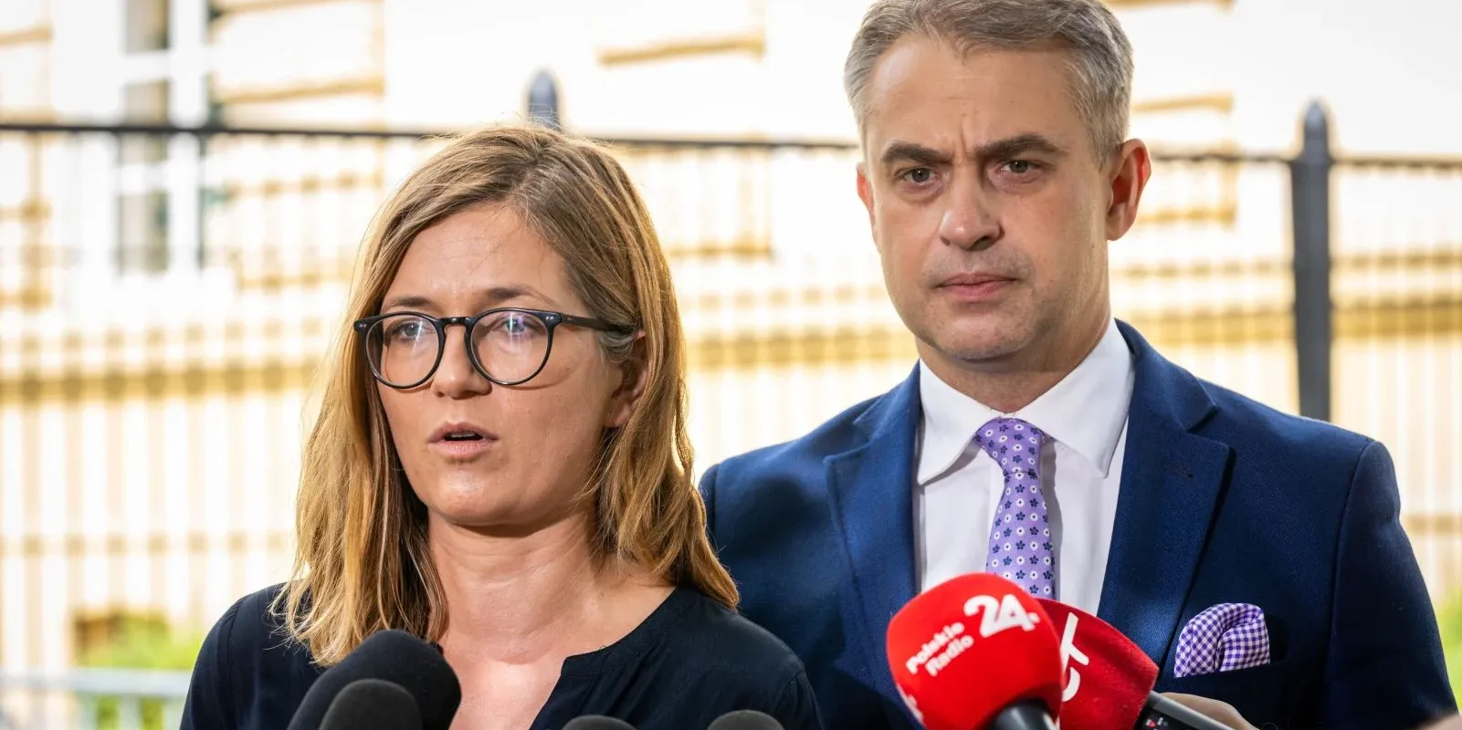 Dwoje polityków przemawia do mediów podczas konferencji prasowej, kobieta z okularami po lewej, mężczyzna w granatowym garniturze po prawej, przed licznymi mikrofonami; w tle ogrodzenie.