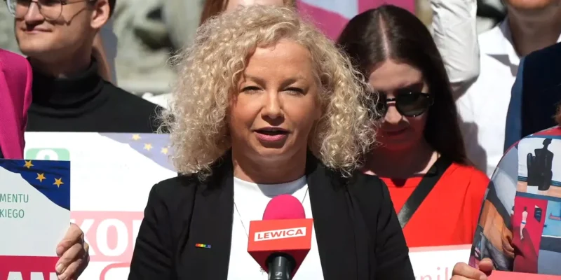 Kobieta o kręconych blond włosach przemawia do mikrofonu z napisem LEWICA podczas wiecu, wokół stoją uczestnicy z plakatami.