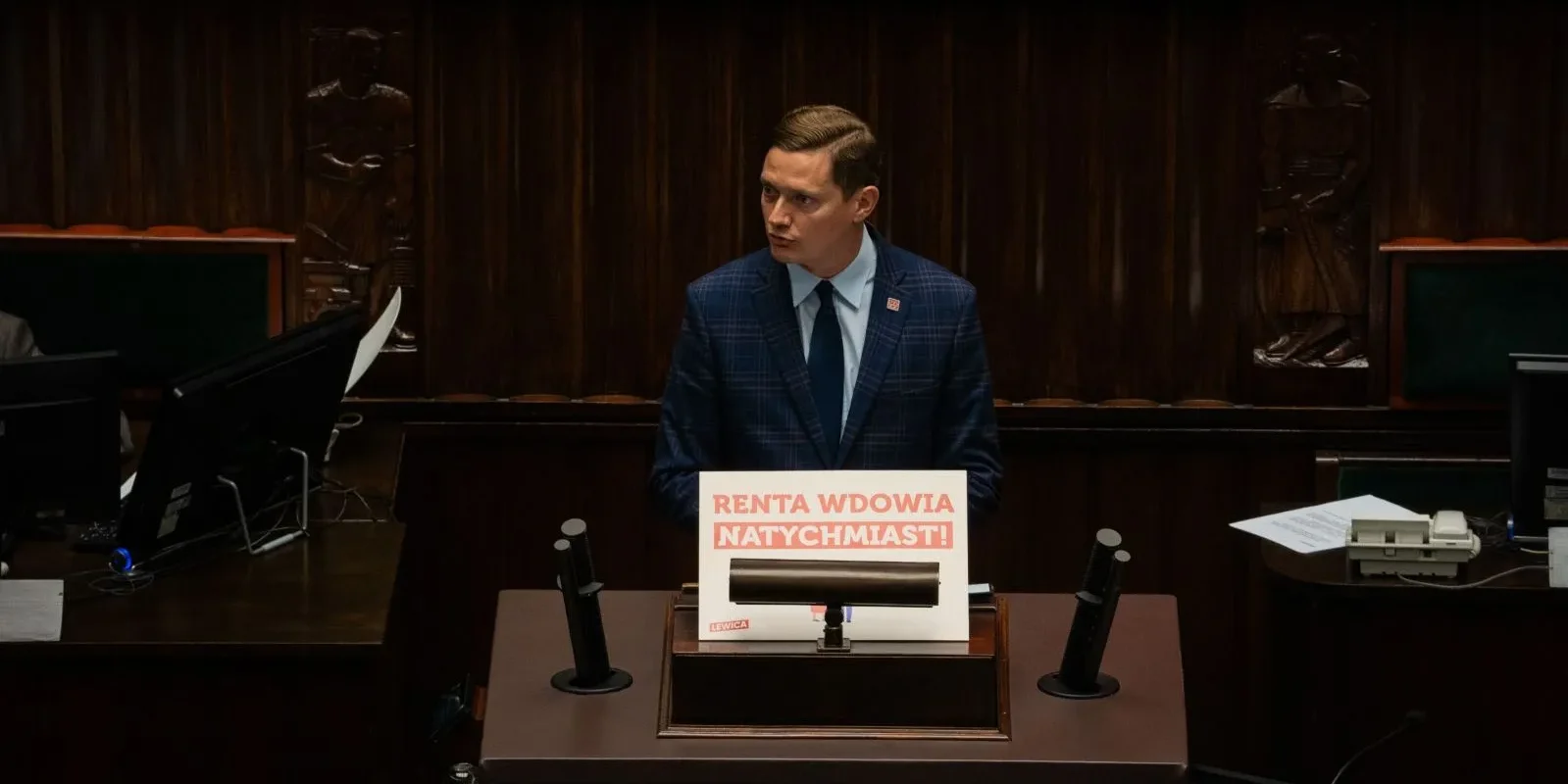 Mężczyzna w garniturze stoi przy mównicy w sali sejmowej, trzymając plakat z napisem „Renta wdowia natychmiast!”.