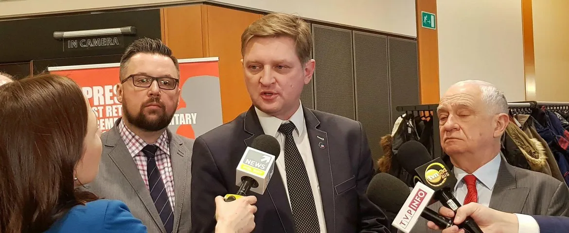 Mężczyzna w garniturze udziela wywiadu grupie dziennikarzy trzymających mikrofony, a obok stoją dwaj inni mężczyźni.