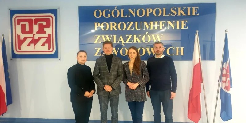 Cztery osoby pozują na tle banera Ogólnopolskiego Porozumienia Związków Zawodowych, z flagami Polski i Unii Europejskiej.