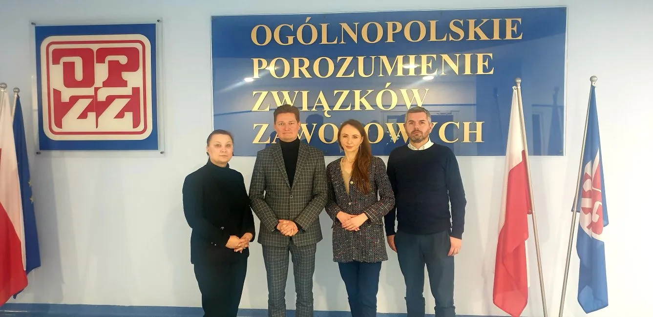 Cztery osoby pozują na tle banera Ogólnopolskiego Porozumienia Związków Zawodowych, z flagami Polski i Unii Europejskiej.