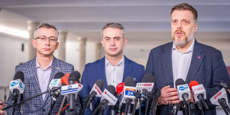 Na zdjęciu trzech mężczyzn w garniturach stoi przed mikrofonami na konferencji prasowej.