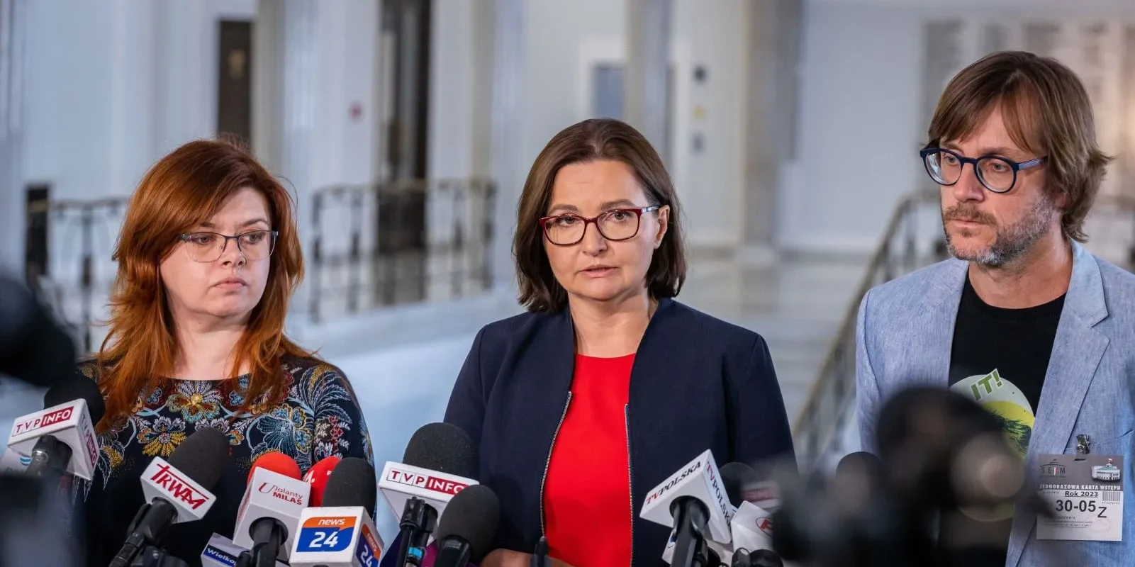 Trzy osoby stojące na konferencji prasowej w jasnym holu, przed licznymi mikrofonami ustawionymi na stanowiskach.