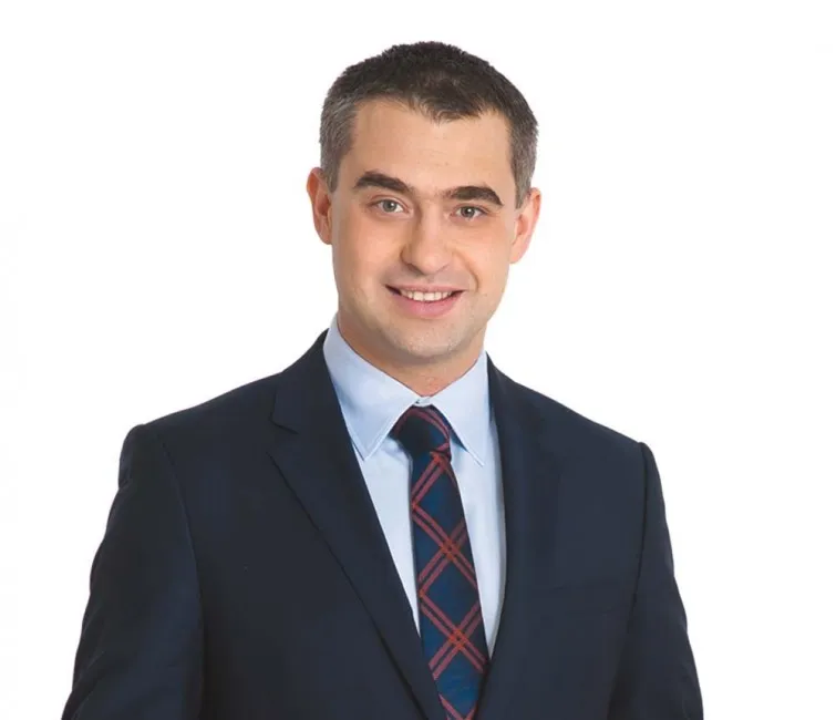 Uśmiechnięty mężczyzna w ciemnym garniturze i kraciastej krawacie na białym tle.