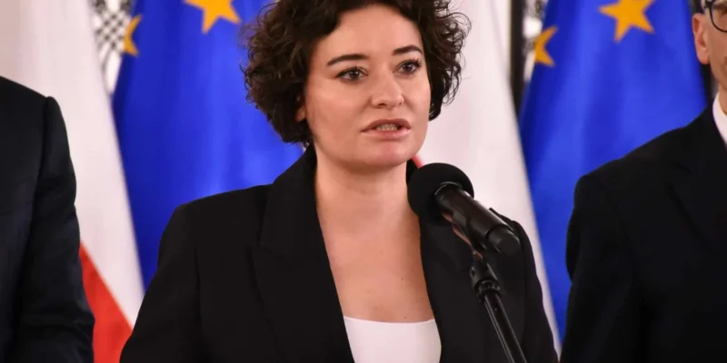 Kobieta w czarnym garniturze przemawia do mikrofonu podczas konferencji, na tle flag Unii Europejskiej.