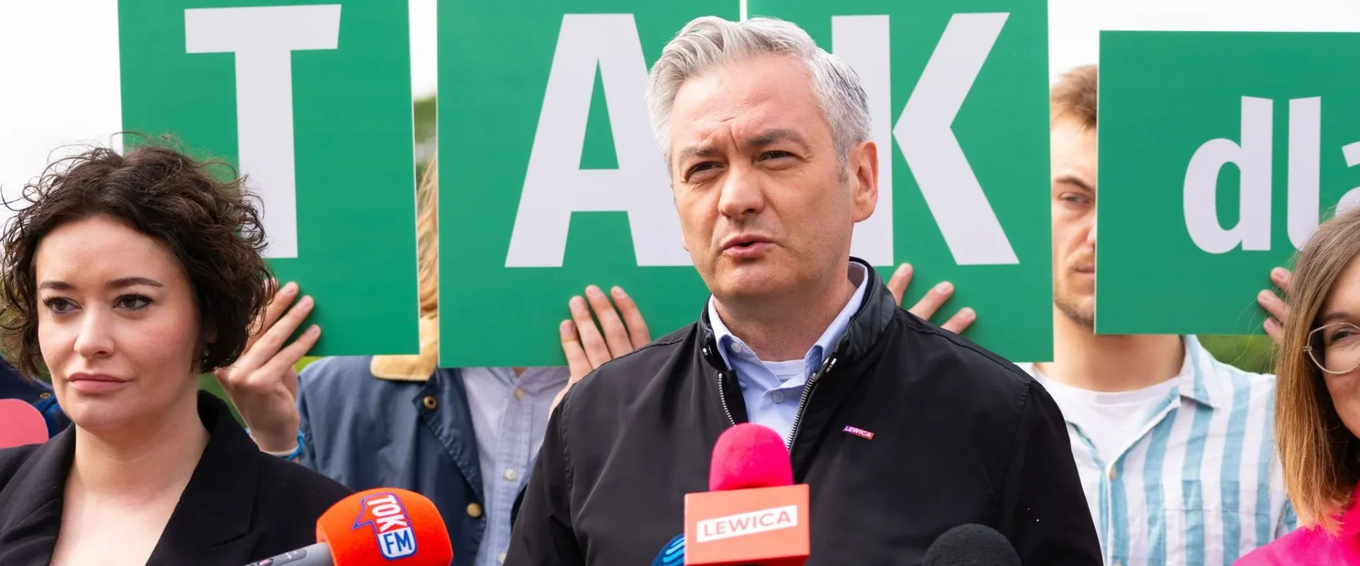 Mężczyzna z siwymi włosami przemawia do mediów podczas konferencji prasowej w kontekście rekonstrukcji rządu, stojąc przed zielonymi tablicami z literami i otoczony reporterami z mikrofonami.