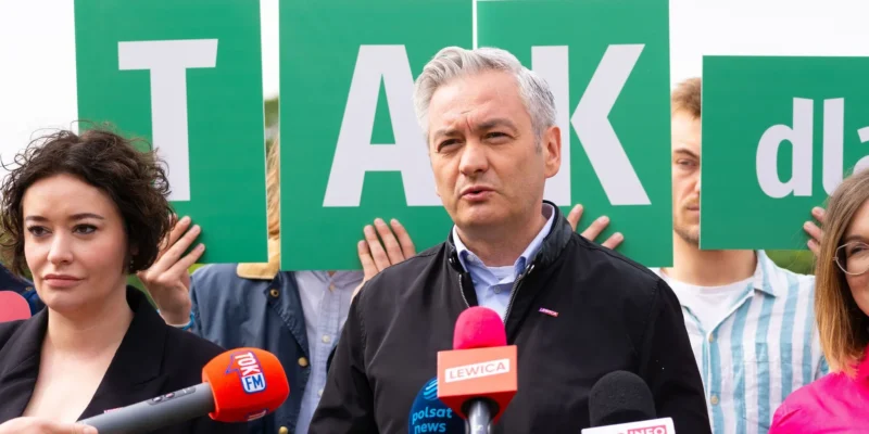 Mężczyzna z siwymi włosami przemawia do mediów podczas konferencji prasowej w kontekście rekonstrukcji rządu, stojąc przed zielonymi tablicami z literami i otoczony reporterami z mikrofonami.