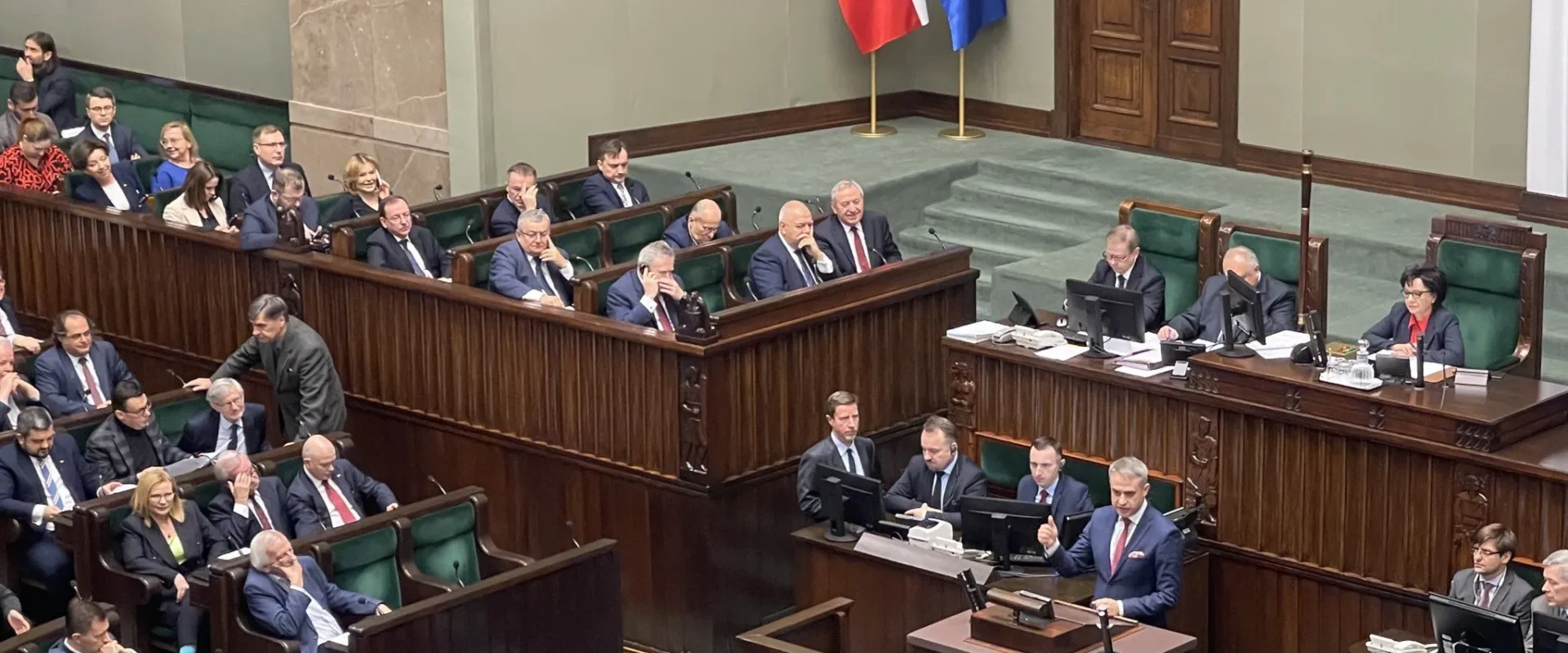 Zdjęcie przedstawia posiedzenie w polskim parlamencie, na którym mówca prezentuje swoje wystąpienie przed zgromadzonymi posłami.