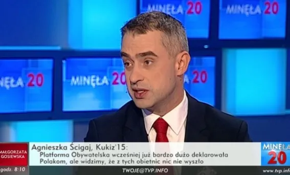 Mężczyzna w garniturze i czerwonym krawacie przemawia w studiu telewizyjnym programu "Minęła 20", z paskiem informacyjnym widocznym u dołu ekranu.