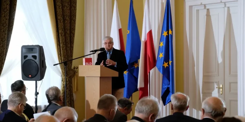 Mężczyzna przemawia przy mównicy przed zgromadzoną publicznością, a w tle widoczne są flagi Polski i Unii Europejskiej podczas konferencji z okazji 20‑lecia Konstytucji.