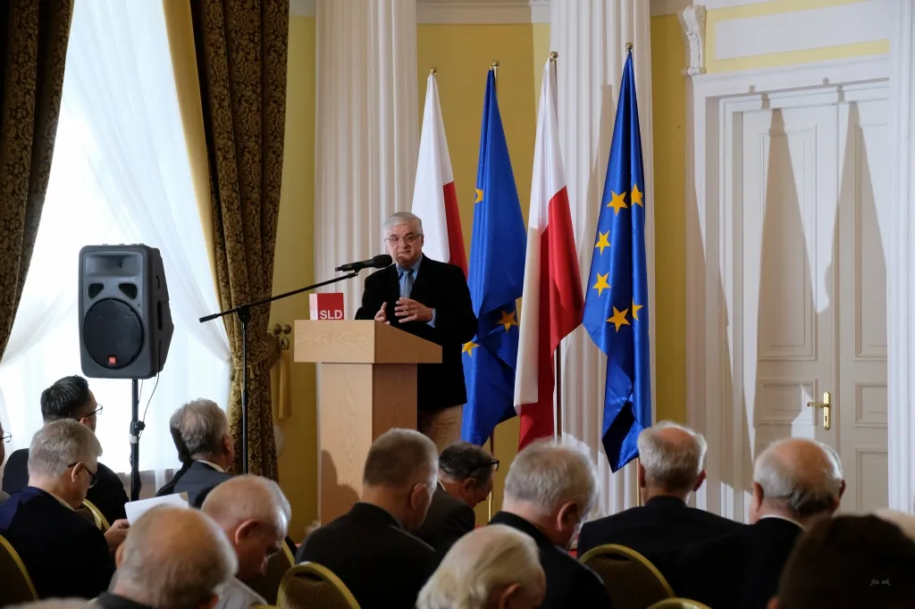 Mężczyzna przemawia przy mównicy przed zgromadzoną publicznością, a w tle widoczne są flagi Polski i Unii Europejskiej podczas konferencji z okazji 20‑lecia Konstytucji.