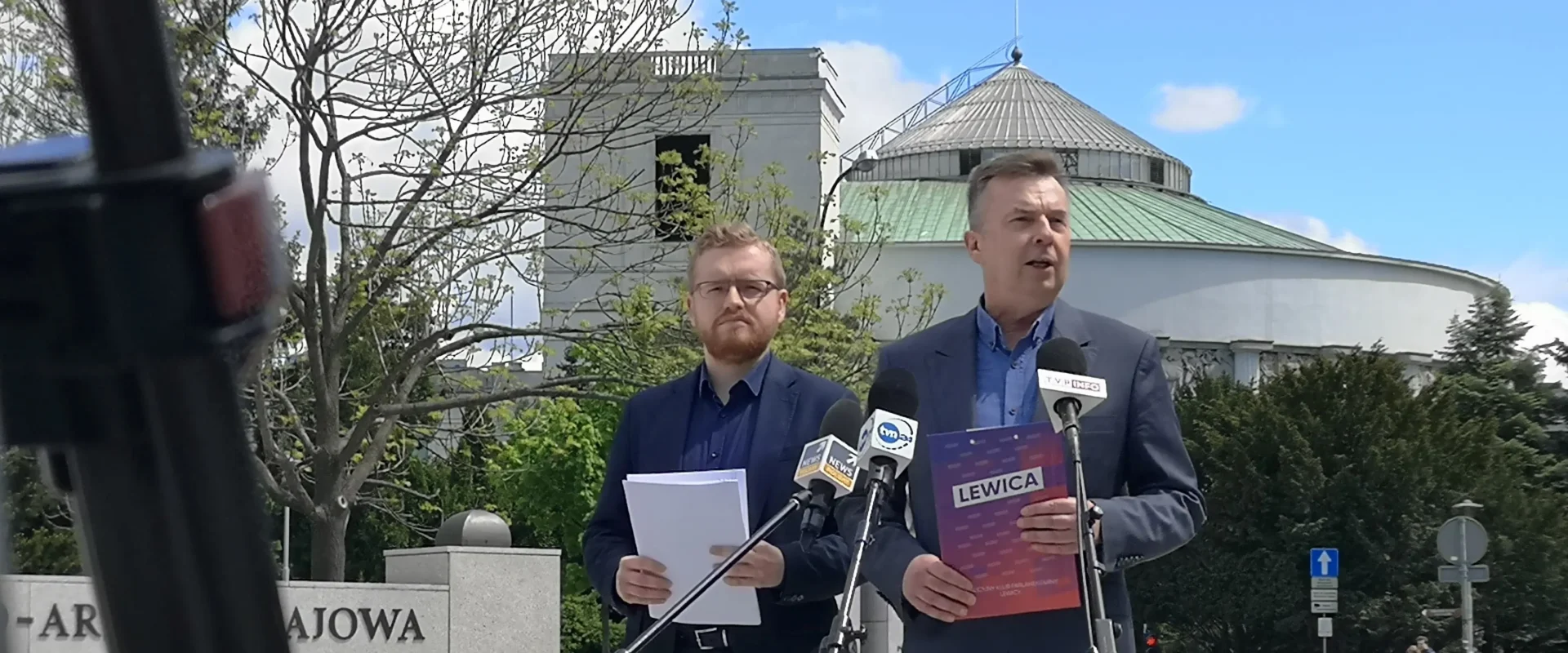 Dwóch mężczyzn w garniturach prowadzi konferencję prasową podczas Festiwalu obietnic PiS na zewnątrz, stojąc przy mikrofonach, z budynkiem z kopułą i polską flagą w tle.