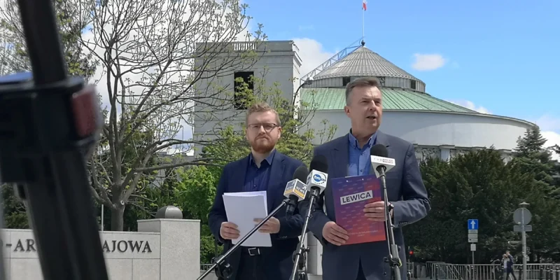 Dwóch mężczyzn w garniturach prowadzi konferencję prasową podczas Festiwalu obietnic PiS na zewnątrz, stojąc przy mikrofonach, z budynkiem z kopułą i polską flagą w tle.