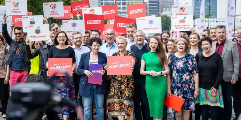 Grupa osób na plenerowym wydarzeniu inaugurującym Wakacyjną Trasę Lewicy, trzyma plakaty z logo Lewica i hasłami.