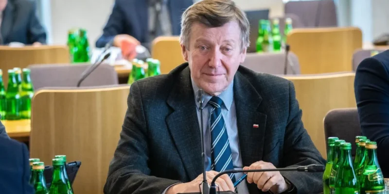 Mężczyzna w garniturze siedzi przy stole w sali konferencyjnej, otoczony zielonymi butelkami wody.