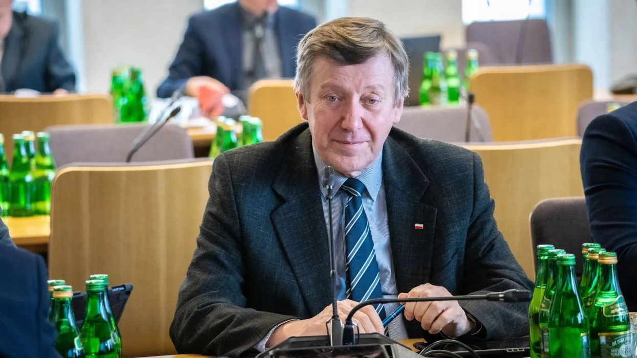 Mężczyzna w garniturze siedzi przy stole w sali konferencyjnej, otoczony zielonymi butelkami wody.