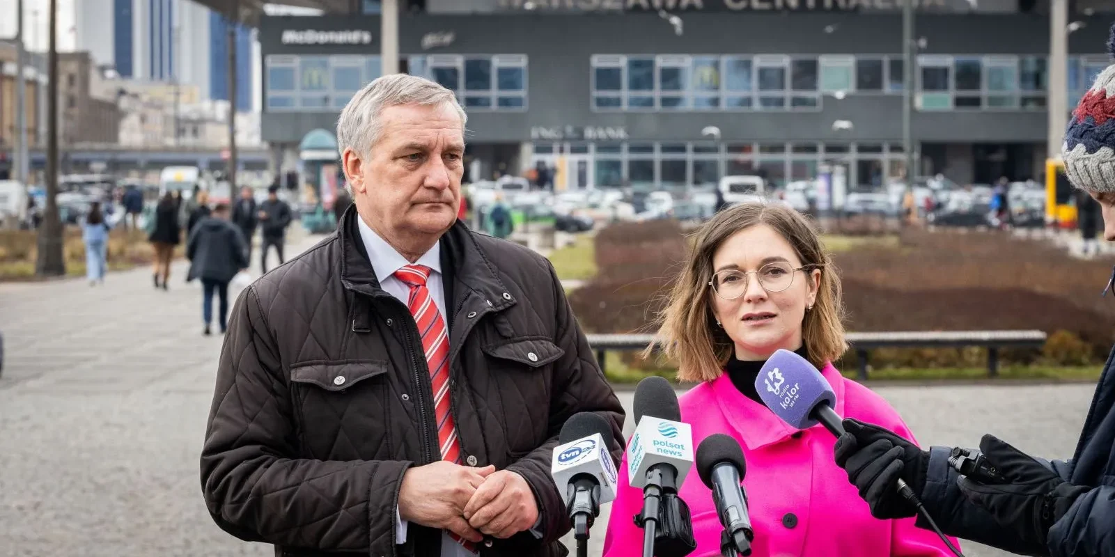 Na zdjęciu widoczna jest dwójka ludzi przemawiających do dziennikarzy przed Warszawą Centralną, a w tle widać ruch uliczny i architekturę miasta.