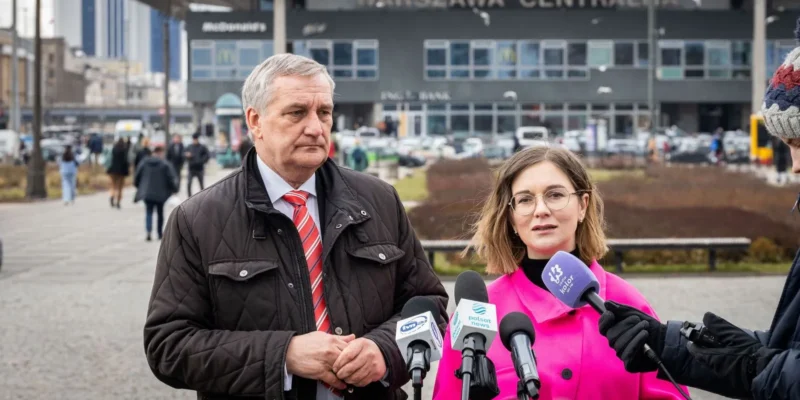 Na zdjęciu widoczna jest dwójka ludzi przemawiających do dziennikarzy przed Warszawą Centralną, a w tle widać ruch uliczny i architekturę miasta.