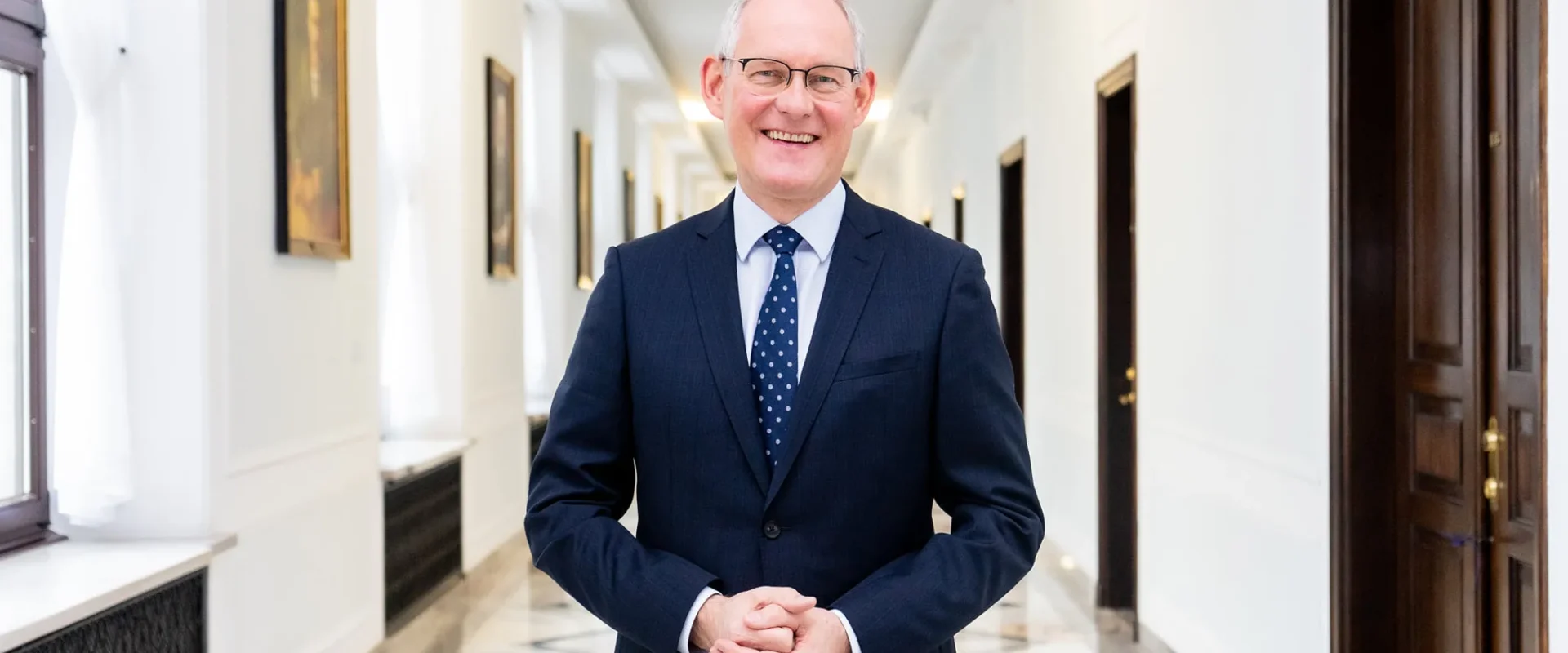 Uśmiechnięty mężczyzna w granatowym garniturze i krawacie z białymi kropkami stoi w jasnym, eleganckim korytarzu z marmurową podłogą i obrazami na ścianach.