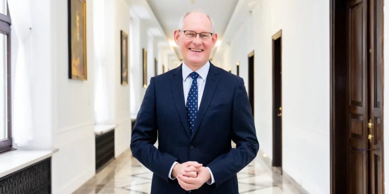 Uśmiechnięty mężczyzna w granatowym garniturze i krawacie z białymi kropkami stoi w jasnym, eleganckim korytarzu z marmurową podłogą i obrazami na ścianach.