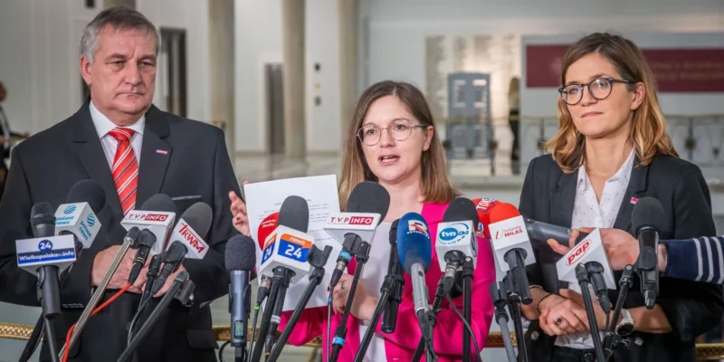 Trzy osoby prowadzą konferencję prasową na temat propozycji Lewicy dotyczącej zerowego VAT-u na bilety kolejowe, stojące przed licznymi mikrofonami.