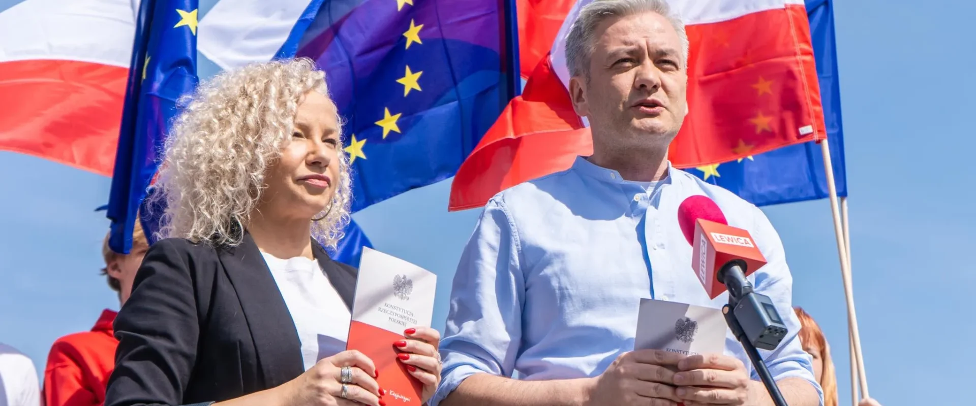 Dwóch polityków stoi przed flagami UE i Polski; kobieta o kręconych blond włosach i mężczyzna w jasnoniebieskiej koszuli przemawiają do kamery, trzymając broszury z wizerunkiem Konstytucji RP.
