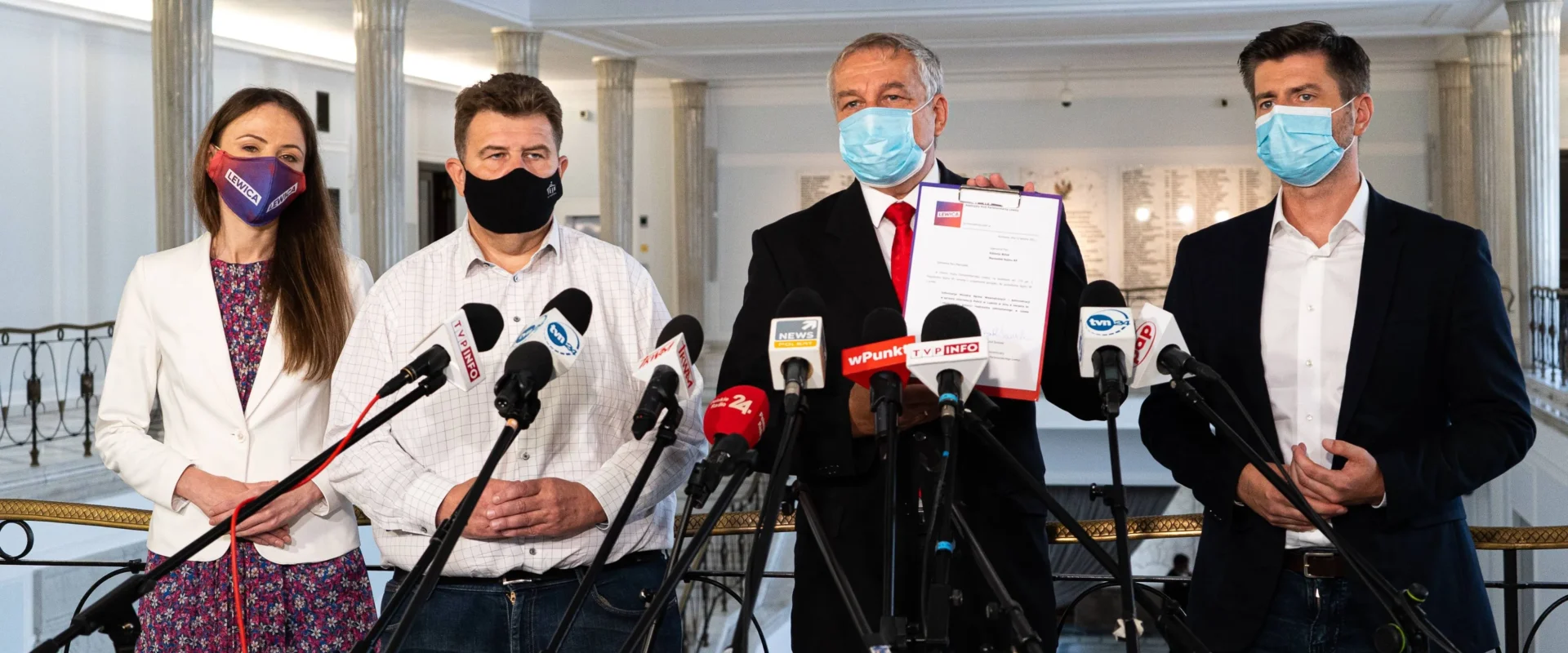 Cztery osoby w maseczkach stoją na konferencji prasowej przy licznych mikrofonach, z czego jedna trzyma dokument.