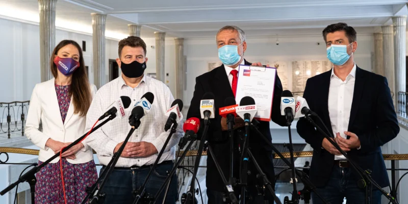 Cztery osoby w maseczkach stoją na konferencji prasowej przy licznych mikrofonach, z czego jedna trzyma dokument.