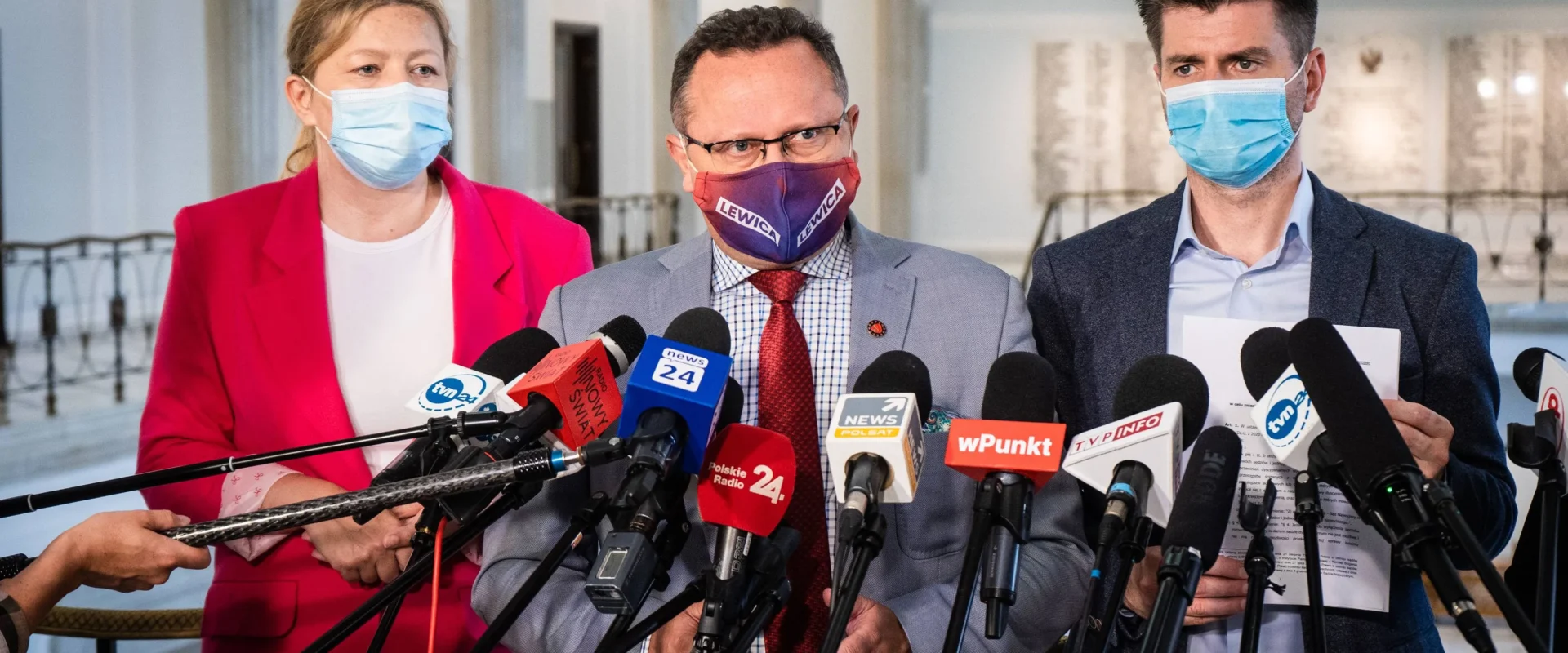 Trzy osoby w maseczkach stoją przed licznymi mikrofonami podczas konferencji prasowej, a środkowy mężczyzna ma maseczkę z napisem Lewica.