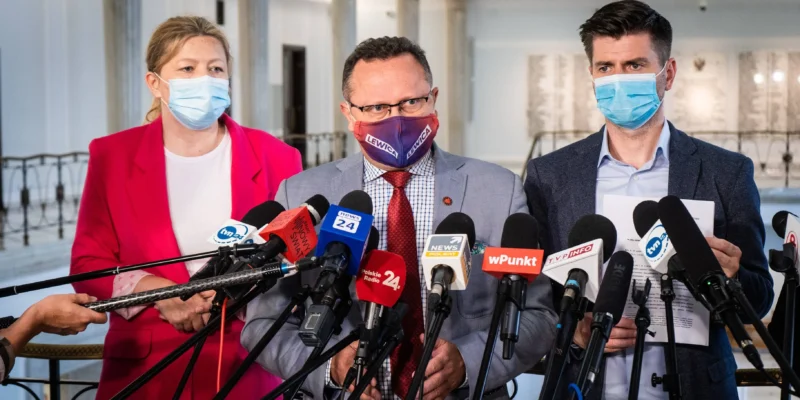 Trzy osoby w maseczkach stoją przed licznymi mikrofonami podczas konferencji prasowej, a środkowy mężczyzna ma maseczkę z napisem Lewica.