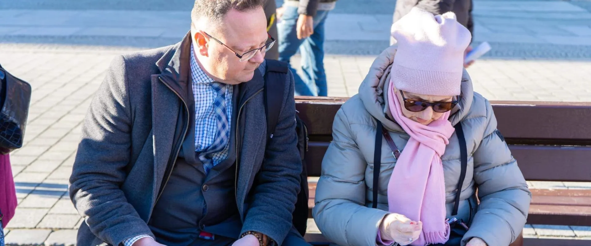 Para siedzi na ławce na placu w Lubelskiem; mężczyzna w granatowym płaszczu przegląda ulotkę, kobieta w różowej czapce i szalu czyta dokumenty.