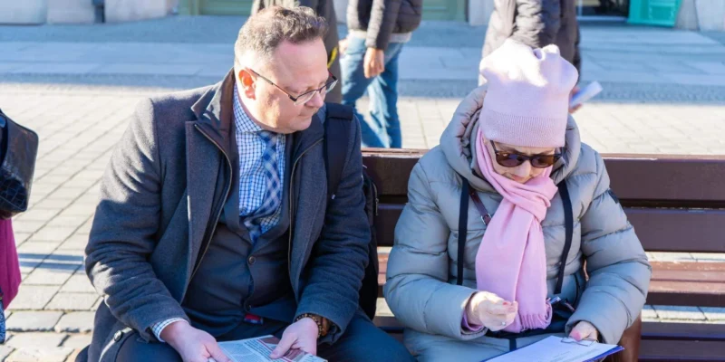 Para siedzi na ławce na placu w Lubelskiem; mężczyzna w granatowym płaszczu przegląda ulotkę, kobieta w różowej czapce i szalu czyta dokumenty.