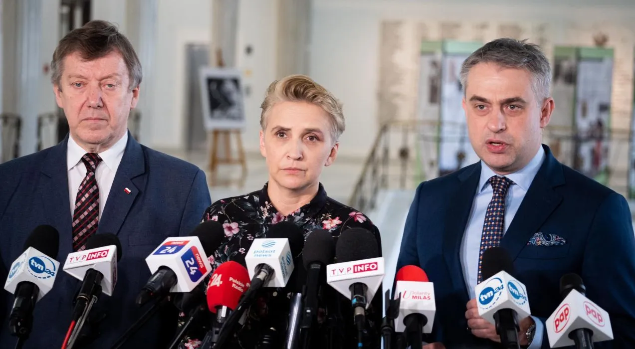 Na zdjęciu trójka polityków udziela wywiadu przed mikrofonami w formalnym otoczeniu.