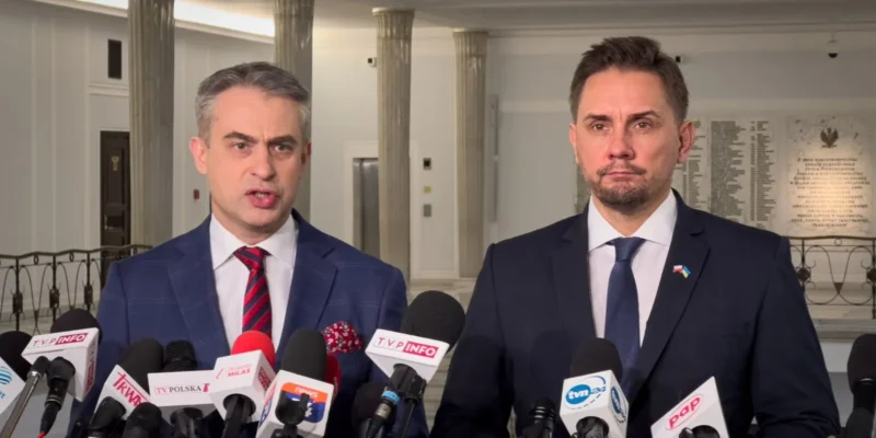 Dwóch mężczyzn w garniturach stoi w holu z kolumnami i udziela konferencji prasowej przed zestawem mikrofonów mediów.