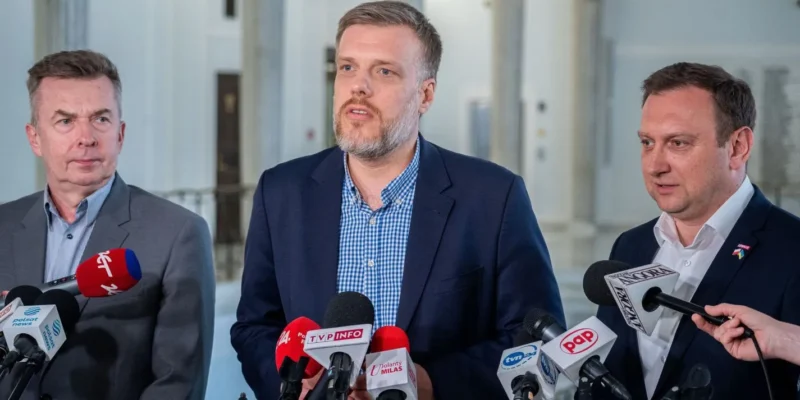 Na zdjęciu trzej mężczyźni stoją przed mikrofonami, prowadząc konferencję prasową w przestronnej, jasnej sali.