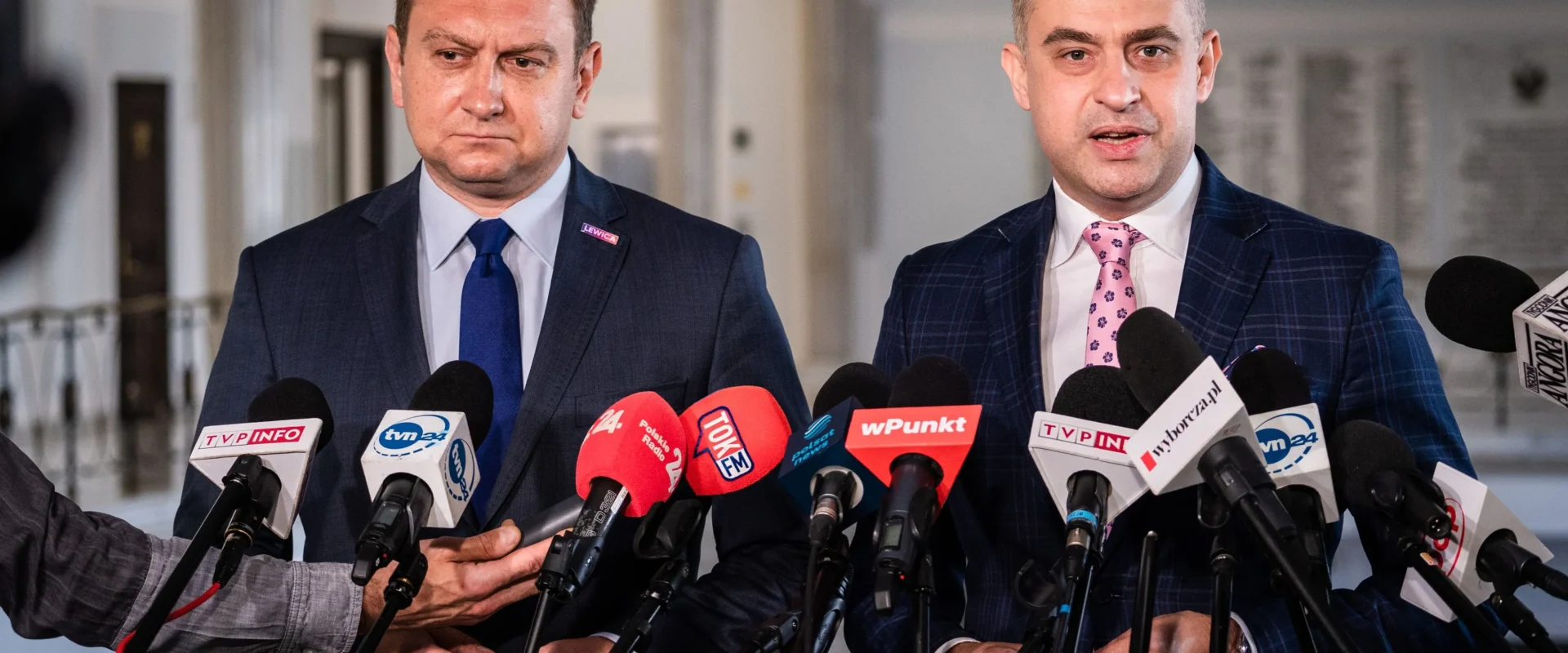 Dwóch mężczyzn w garniturach stoi przy licznych mikrofonach podczas konferencji prasowej w korytarzu.