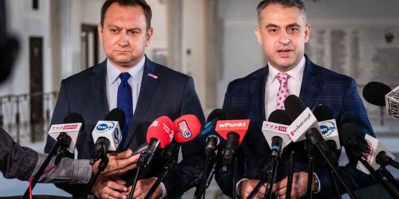 Dwóch mężczyzn w garniturach stoi przy licznych mikrofonach podczas konferencji prasowej w korytarzu.