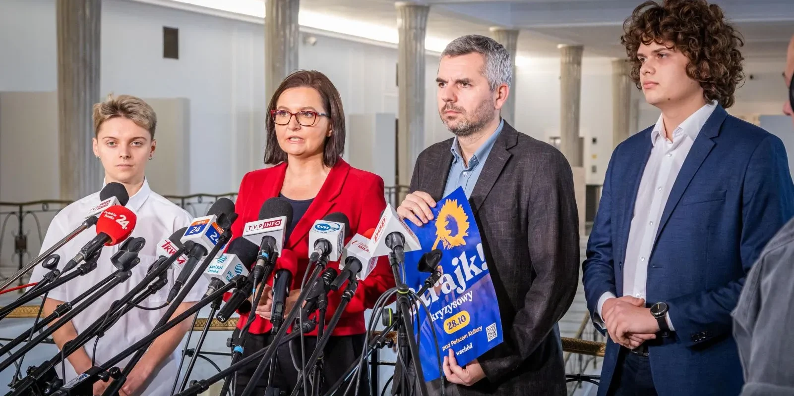 Na zdjęciu czterech ludzi stoi przed mikrofonami, z jedną osobą w czerwonym ubraniu wygłaszającą przemówienie, a obok niej trzej mężczyźni, w tym jeden trzymający plakat zapraszający na strajk.