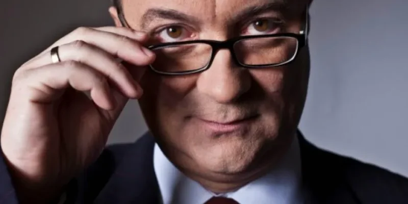 Zbliżenie na mężczyznę w garniturze i krawacie poprawiającego okulary i patrzącego w obiektyw podczas rozmowy.