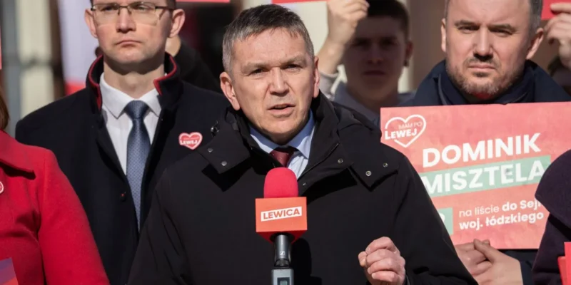 Mężczyzna w ciemnym ubraniu przemawia do kamery podczas wiecu Lewicy, trzymając czerwony mikrofon, a w tle widoczni są ludzie oraz plakaty wyborcze.