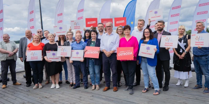 Grupa uczestników marszu 1 października stoi na drewnianym pomoście nad wodą, trzymając plakaty z hasłami Lewica, Prawa Kobiet i Świeckie państwo.