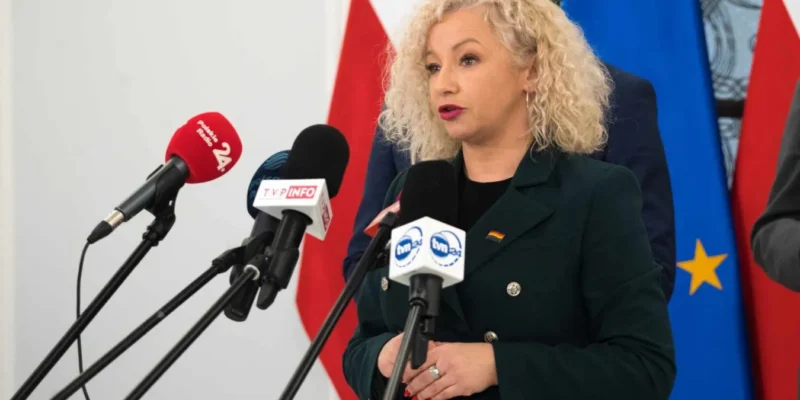 Polityczka w zielonej marynarce przemawia na konferencji prasowej, stojąc przy mikrofonach, z flagami państwowymi w tle.